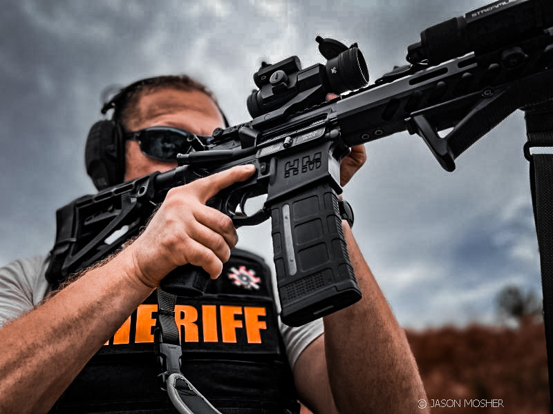 A person shooting an AR-15 with a black vest on that says "Sheriff". 