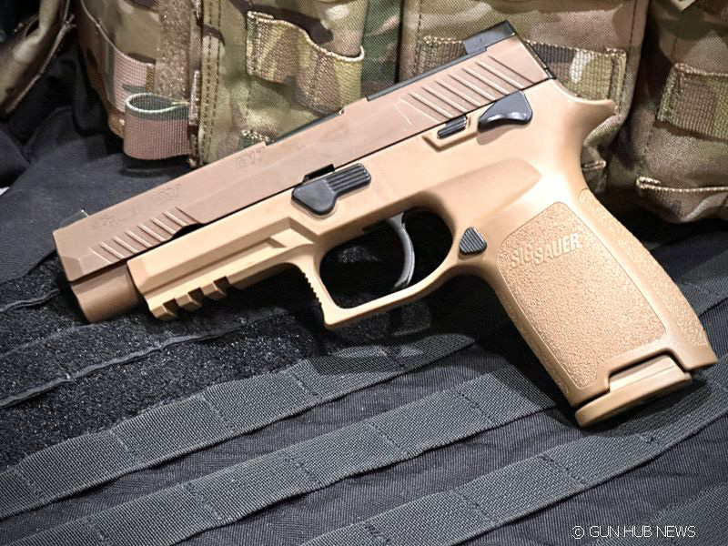 A brown gun on a black table with a camo vest in the background. 