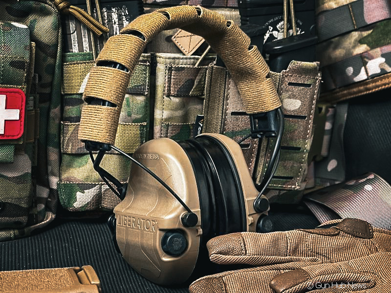 A pair of brown hearing protection earmuffs in front of a camo vest and pair of gloves.